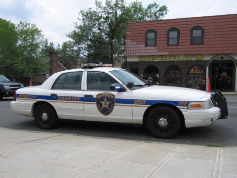 1991 Ford LTD Crown Victoria - Driver-side profile of a white Essex Sheriff Ford Crown Victoria with blue-and-gold striping parked along a storefront street.