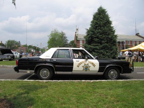1991 Ford LTD Crown Victoria - Black-and-white Highway Patrol sedan shown in profile beside a row of evergreens and event tents.