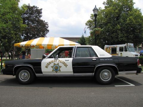 1991 Ford LTD Crown Victoria - Side profile of a black-and-white Highway Patrol sedan parked beside a yellow-and-white striped tent.