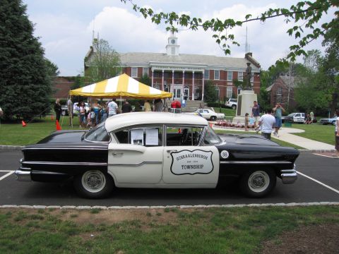 1991 Ford LTD Crown Victoria - Side profile of a black-and-white 1950s police sedan marked Schraalenburgh Township in front of a brick building.