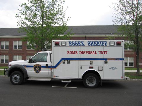 1991 Ford LTD Crown Victoria - Driver-side profile of a white Essex Sheriff Bomb Disposal Unit box truck with blue and orange stripes, parked before a brick building.