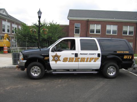 1991 Ford LTD Crown Victoria - Driver-side view of a black-and-white Passaic County Sheriff Mounted Patrol Ford Excursion SUV in a lot beside a brick building.
