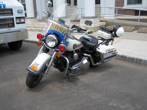 1991 Ford LTD Crown Victoria - white Essex County Sheriff police touring motorcycle with windshield, fairing flag and top case, parked on pavement.
