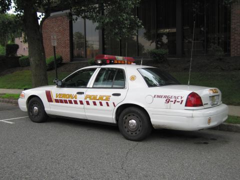 1991 Ford LTD Crown Victoria - Rear driver-side view of a white Verona Police Crown Victoria cruiser with maroon striping, parked at a curb by trees.
