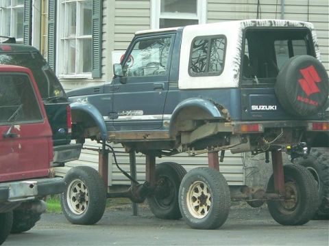 1991 Ford Bronco - A heavily modified blue Suzuki Samurai is parked next to a red Ford Bronco. The Suzuki has been significantly lifted with a custom suspensi…