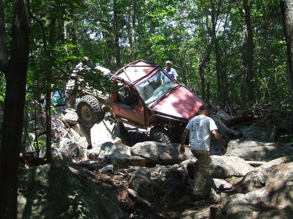 Modified off-road vehicle with roll cage climbing over large rocks, another vehicle visible behind it in trees