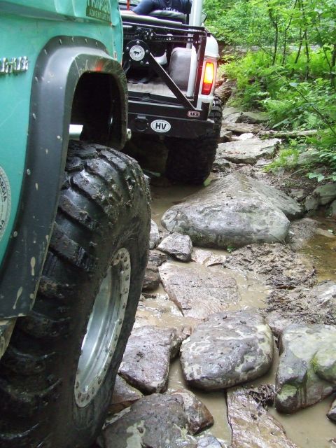 1967 Ford Bronco - Two off-road vehicles tackle a challenging rocky and muddy trail through a wooded area.