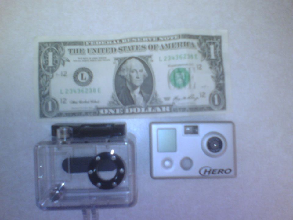 GoPro camera in its case next to a silver GoPro Hero camera and a one dollar bill