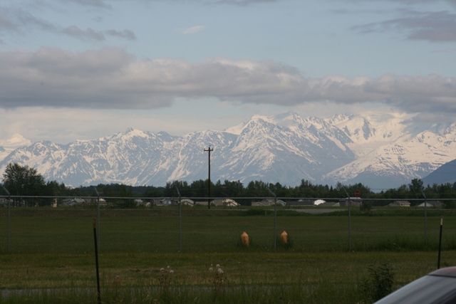 Snow-capped mountains under a cloudy sky, with a fence and grassy field in the foreground