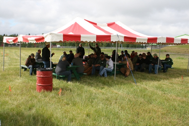 Group of people sitting at picnic tables under a red and white striped tent in a grassy field