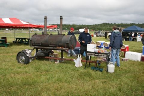 1990 Chevrolet Suburban - An outdoor gathering features a large, trailer-mounted barbecue smoker, with people mingling and other vehicles visible in the distance.