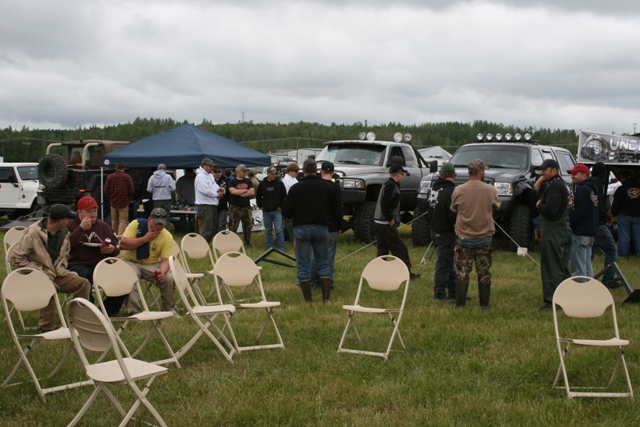 People standing and sitting in chairs around several large, modified pickup trucks outdoors