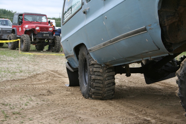 Light blue SUV with large off-road tires, seen from the rear quarter, with a red Jeep in the background