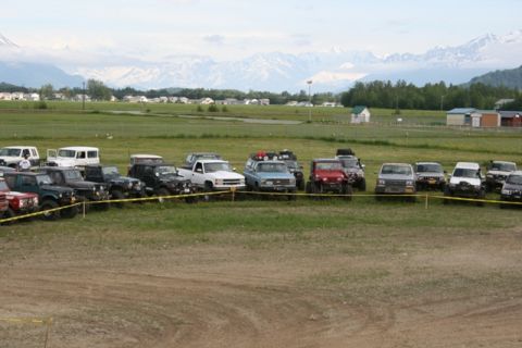 1990 Chevrolet Suburban - A variety of SUVs and trucks are parked in a line on a grassy field, with mountains visible in the distance.