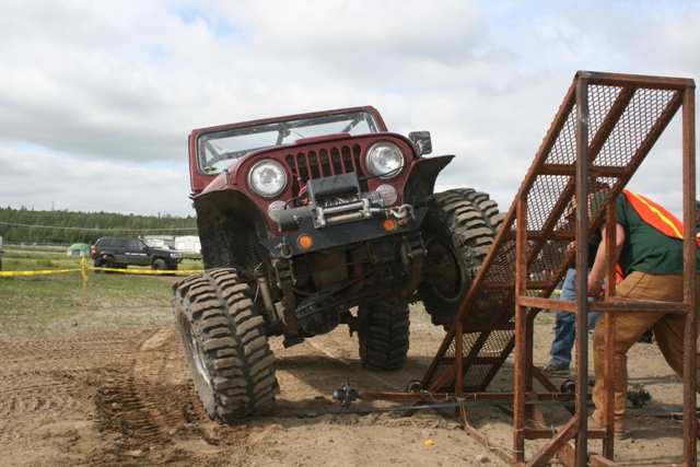 Red Jeep CJ-7 with large tires climbing a metal ramp, another SUV in background