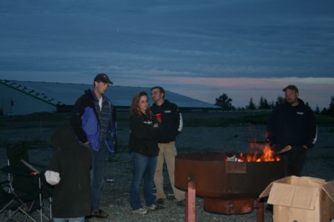 1990 Chevrolet Suburban - A group of people gather around a fire pit as the sky darkens, with a large structure visible in the distance.