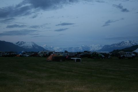 1990 Chevrolet Suburban - A gathering of vehicles and tents is set up in a grassy field, with a mountain range visible in the distance under a cloudy sky.