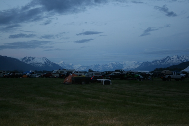 Field with multiple vehicles and tents, snow-capped mountains in background