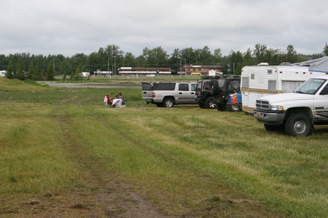 Silver SUV with open hatch, black Jeep, white pickup truck, and a camper trailer parked in a grassy area with people sitting nearby
