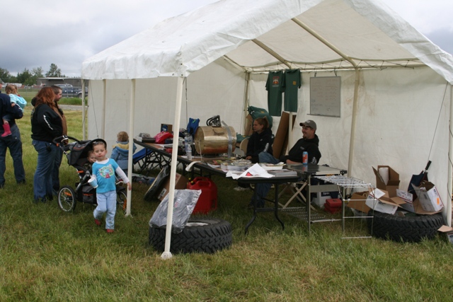 People gathered under a white tent at an outdoor event, with tables displaying items and children playing