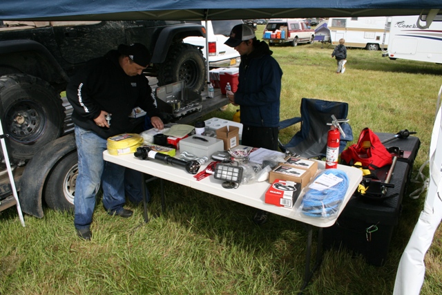 Two men examine car parts on a folding table under a blue canopy, with a large off-road vehicle and trailers in the background