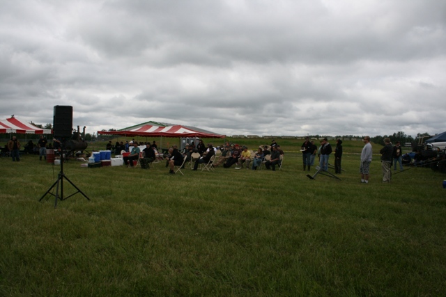 Outdoor gathering with red and white striped tents, people sitting and standing, and a speaker on a stand
