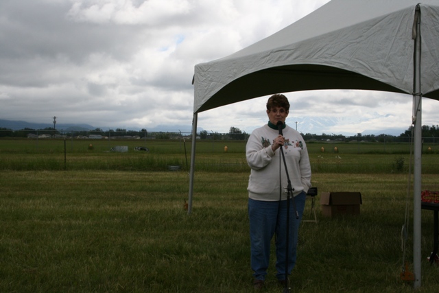 Woman holding a microphone stands under a white tent in a grassy field with mountains in the distance