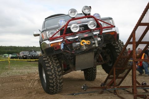 1990 Chevrolet Suburban - A heavily modified black SUV with large off-road tires and a red bull bar with multiple spotlights and a winch is positioned on a sandy inc…