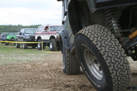 1990 Chevrolet Suburban - A line of off-road vehicles are parked on a dirt track, with the front tire of a dark-colored SUV in the foreground. The vehicles appear to…