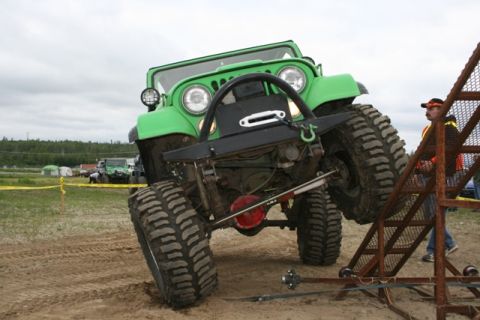 1990 Chevrolet Suburban - A bright green modified Jeep is angled on a dirt track, with its front tires elevated on a metal ramp structure. A man in an orange safety…