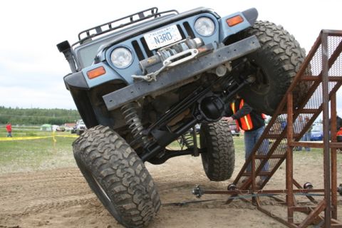 1990 Chevrolet Suburban - A blue Jeep Wrangler is tilted at a steep angle, with its front tire on a metal ramp. The vehicle has large off-road tires and a winch moun…