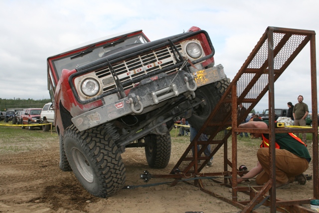 Red Ford Bronco tilted on a metal ramp structure with people around.