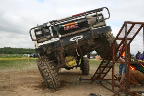 1990 Chevrolet Suburban - A heavily modified white GMC SUV is tilted at a steep angle on a sandy track, with its front tire resting on a metal ramp structure. The ve…