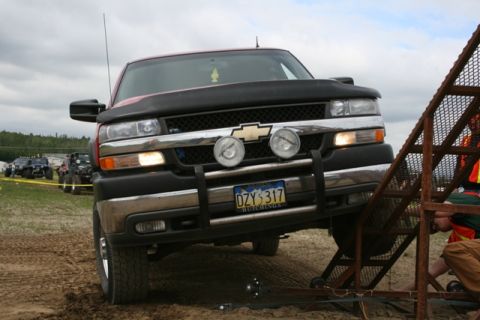 1990 Chevrolet Suburban - A red Chevrolet pickup truck is positioned at an angle on a metal ramp, with its front tires elevated. Other off-road vehicles are visible…