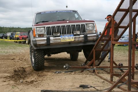 1990 Chevrolet Suburban - A maroon Jeep Cherokee is positioned at the bottom of a metal ramp structure, with its front tires in the dirt. Several other off-road vehi…
