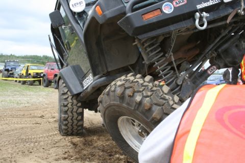 1990 Chevrolet Suburban - A close-up, low-angle shot of the undercarriage and large off-road tires of a dark green Jeep. Several other off-road vehicles are parked i…