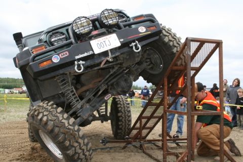1990 Chevrolet Suburban - A heavily modified green Jeep Wrangler is angled precariously on a metal ramp structure, with a man in an orange safety vest kneeling besid…