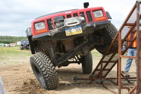 1990 Chevrolet Suburban - A red Jeep Cherokee is angled onto a metal ramp, with its front tire on the ramp and its rear tire on the ground. The vehicle has large off…