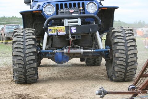 1990 Chevrolet Suburban - The front axle and massive tires of a lifted blue Jeep Wrangler are shown in close-up, with a winch mounted on the front bumper. The vehicl…