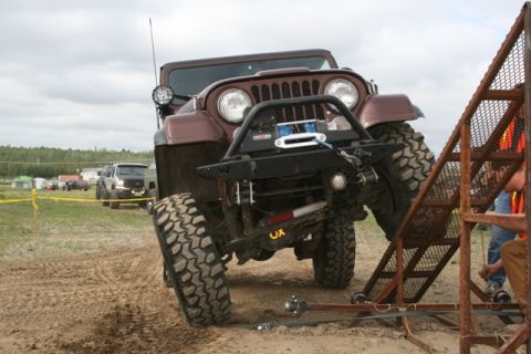 1990 Chevrolet Suburban - A lifted, maroon Jeep Wrangler with large off-road tires is positioned on a dirt track, facing a metal ramp structure. The Jeep has a winch…