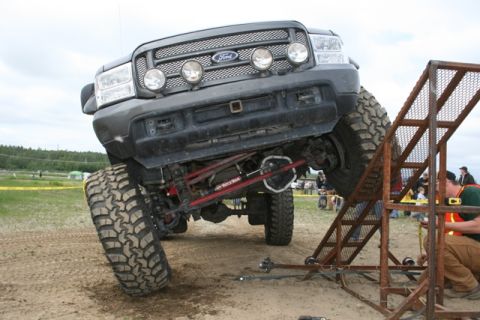 1990 Chevrolet Suburban - A heavily modified dark gray Ford Super Duty truck is at an extreme angle, with its front passenger-side tire on a metal ramp structure. Th…