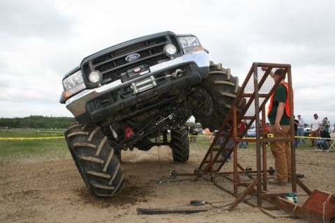 1990 Chevrolet Suburban - A large, modified black Ford truck with oversized tires is tilted at an extreme angle, with its front tire resting on a metal ramp structur…