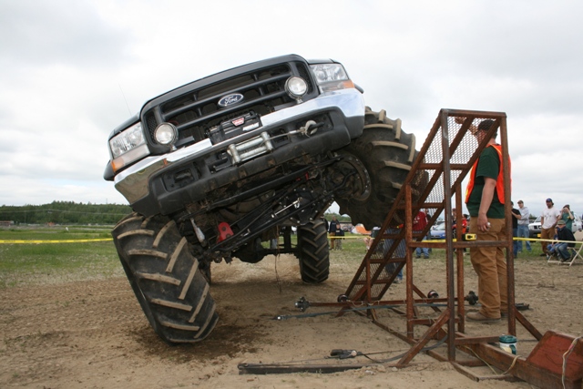 Modified black Ford truck with huge tires tilted on a metal ramp with a person standing on it.