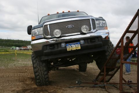 1990 Chevrolet Suburban - A lifted, dark-colored Ford Super Duty pickup truck is angled on a dirt track, with its front tire climbing a metal ramp. The truck has a c…
