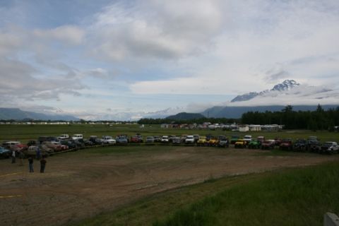 1990 Chevrolet Suburban - A large group of off-road vehicles, including Jeeps and SUVs, are parked in a field with mountains in the background under a cloudy sky.