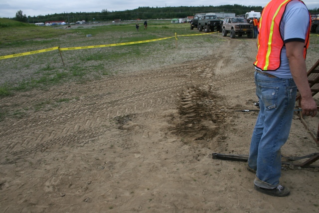 Person in safety vest and jeans near off-road vehicles on a dirt track.