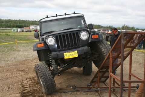 1990 Chevrolet Suburban - A black Jeep Wrangler is angled on a sandy surface, with its front tire climbing a metal ramp. The Jeep has a yellow license plate reading…
