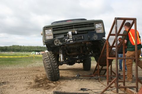 1990 Chevrolet Suburban - A heavily modified, lifted green Chevrolet truck is positioned at an angle on a dirt track, with its front tire resting on a metal ramp str…