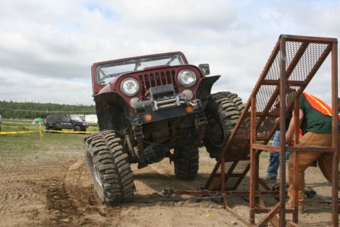1990 Chevrolet Suburban - A lifted, red Jeep CJ is angled on a dirt track, with its front tires climbing a metal ramp. A person in a green shirt and orange vest is v…