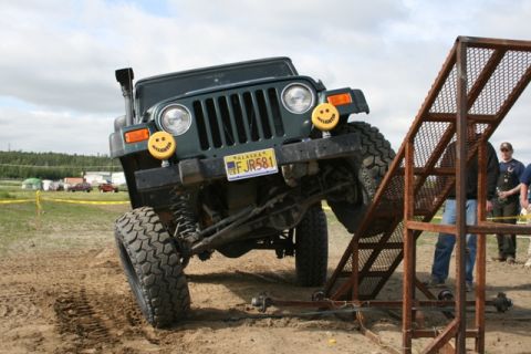 1990 Chevrolet Suburban - A dark green Jeep Wrangler is angled precariously on a metal ramp, with its front tires elevated. The Jeep has two yellow smiley face light…